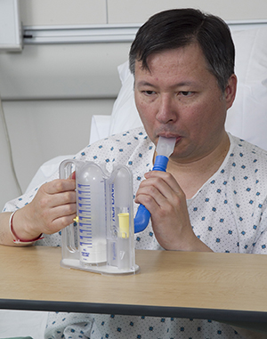 Hombre sentado en la cama de un hospital con el tubo de un espirómetro de incentivo en su boca.