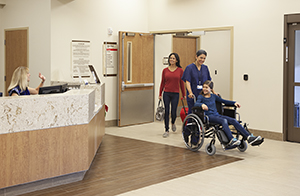 Un proveedor de atención médica llevando a una niña en silla de ruedas hacia la salida del hospital y una mujer caminando a su lado.